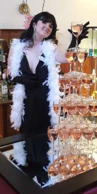 A woman in a cocktail dress standing next to a champagne fountain for Death on the Rocks